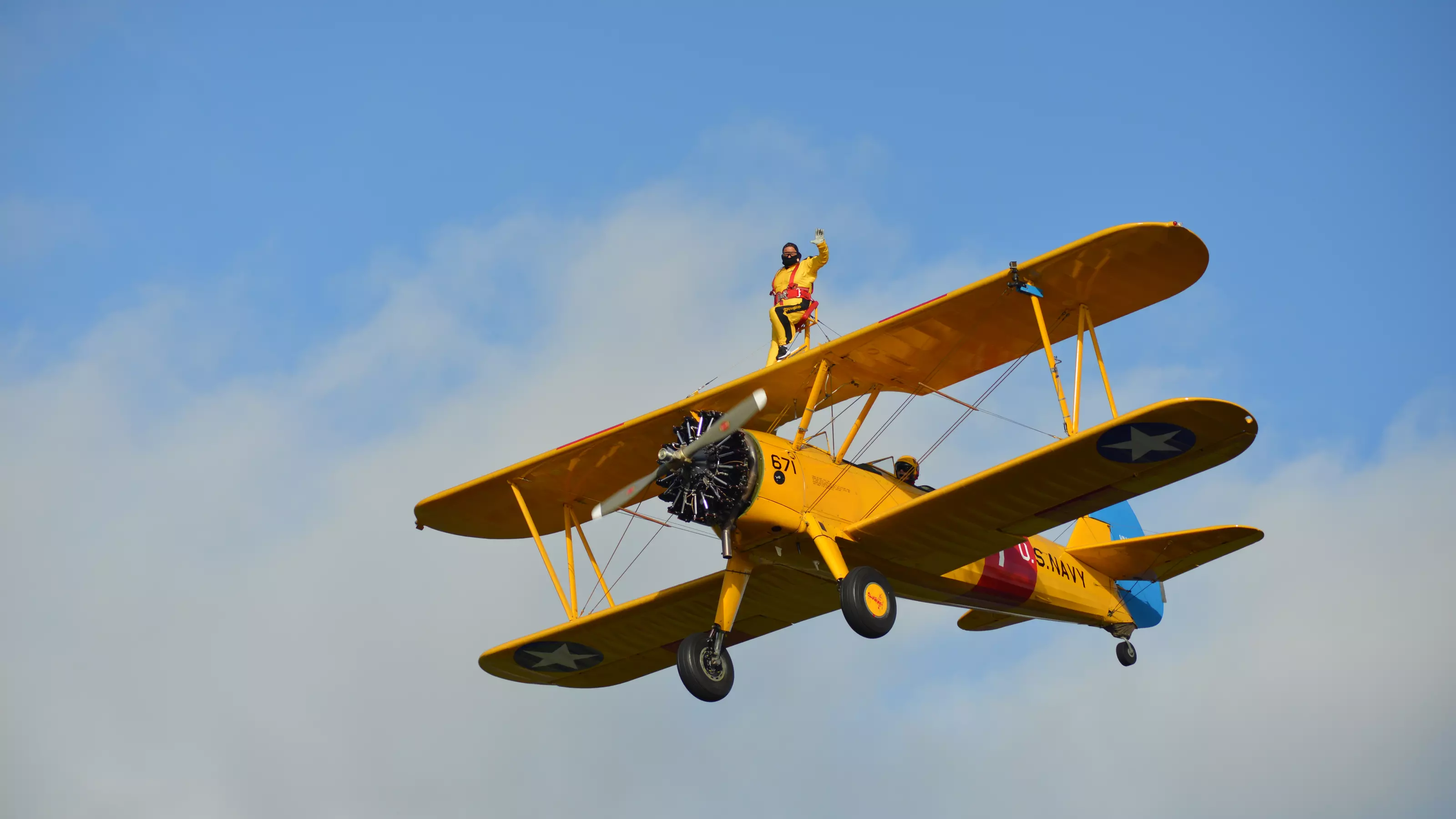 Man waving while safely strapped to the wing of a yellow plane as part of the Wingwalk challenge event.