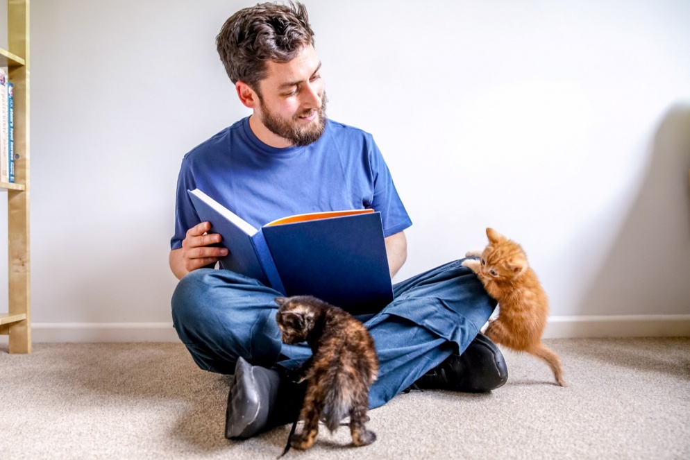 Man sitting on the floor with open book and two kittens climbing on his lap
