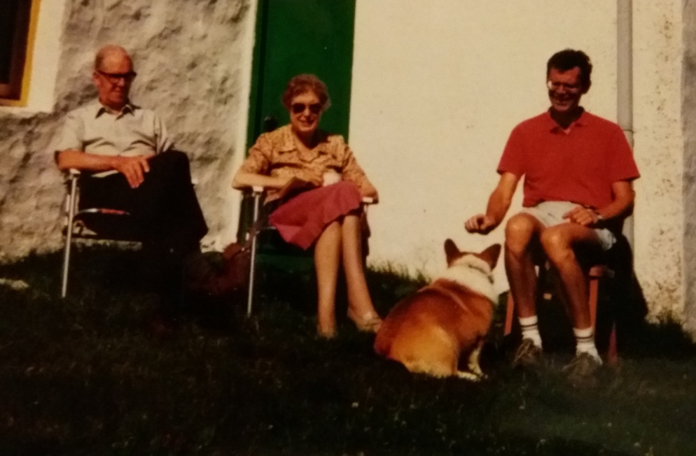 Old photo showing three people sat on chairs with corgi at their feet
