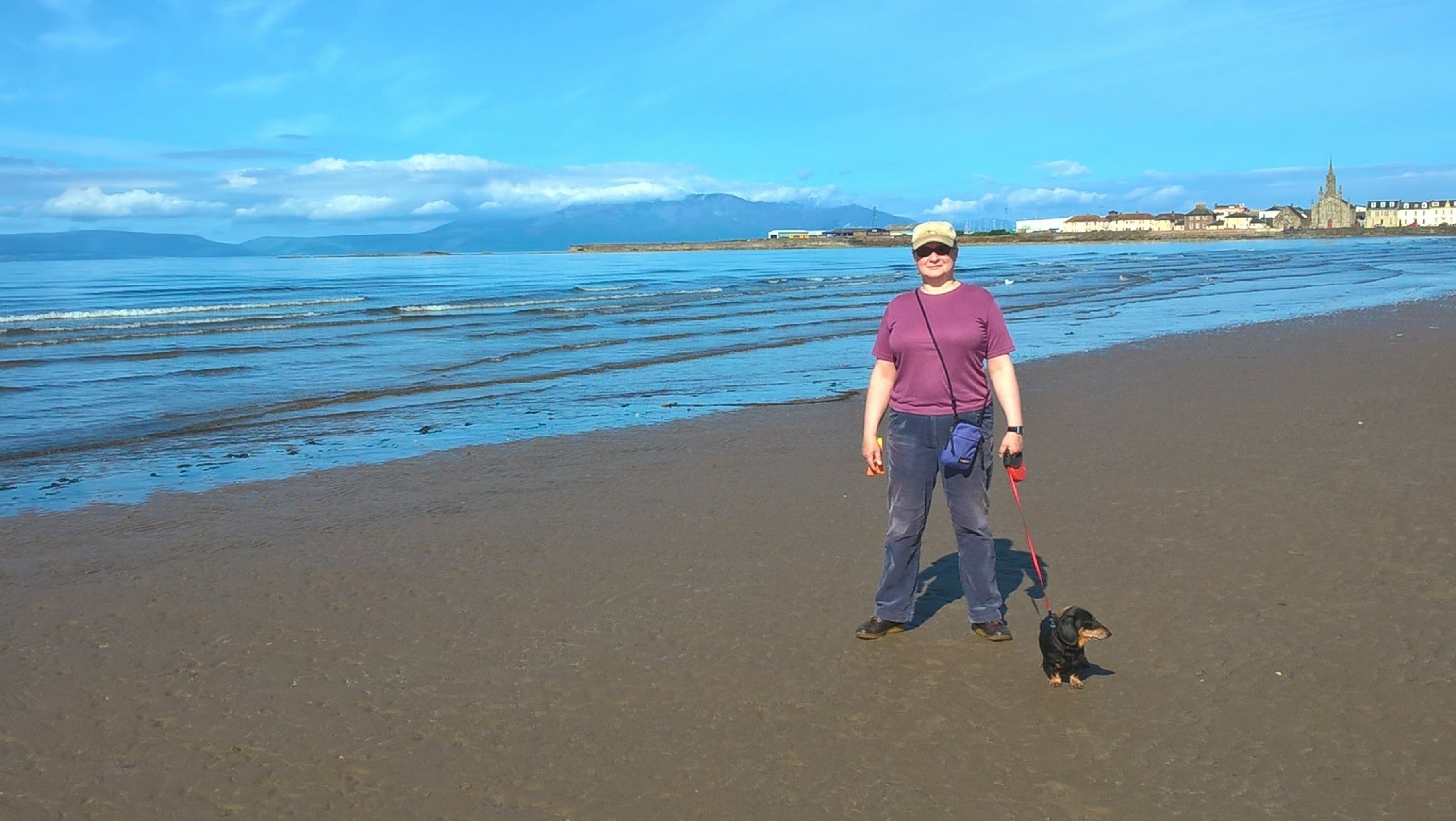 A casually-dressed woman and her black and tan dachshund dog stand on a sandy beach with sea and blue sky in the background