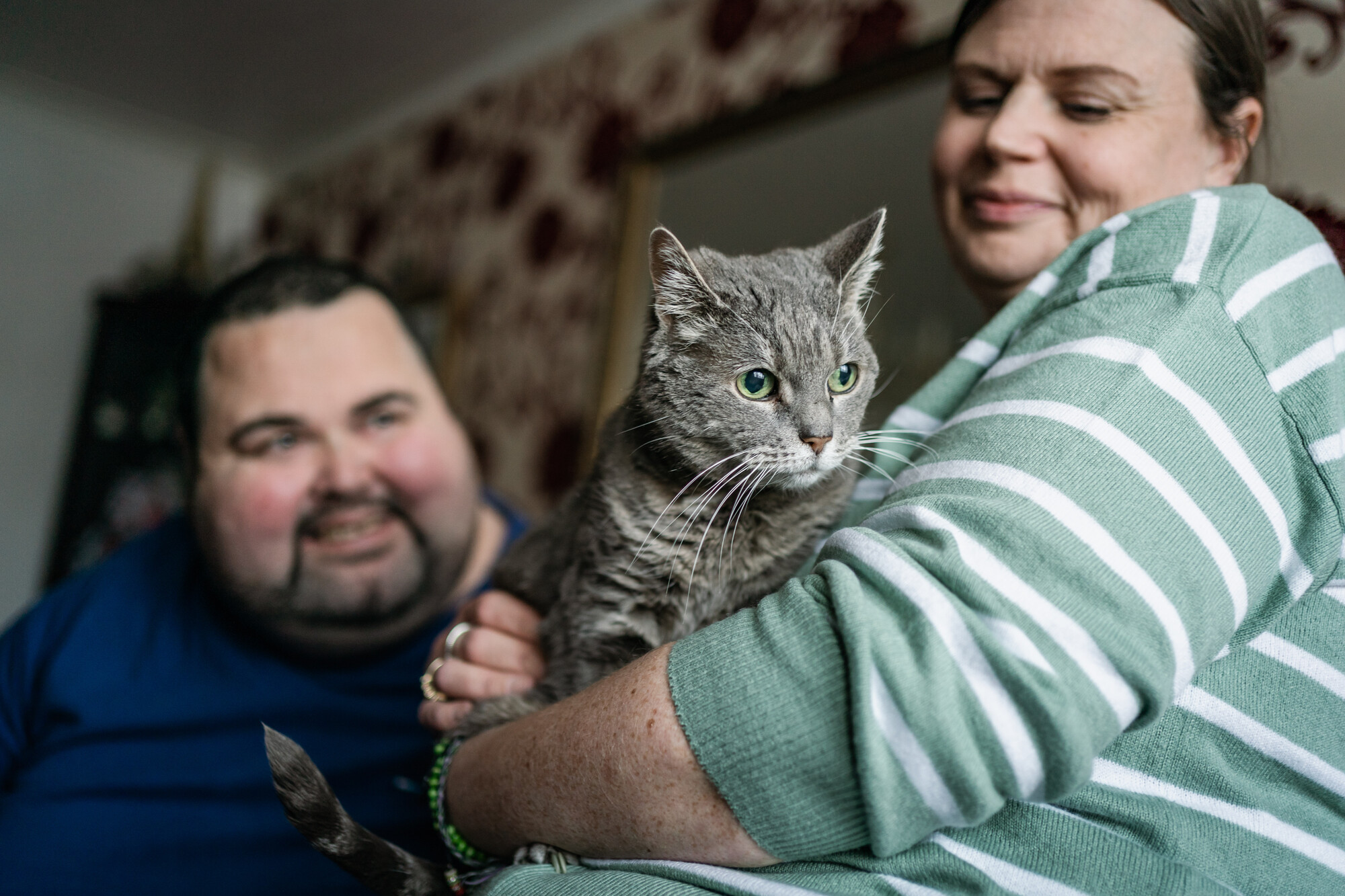 Woman in green and white striped jumper holding grey cat Inari with other owner smiling in background