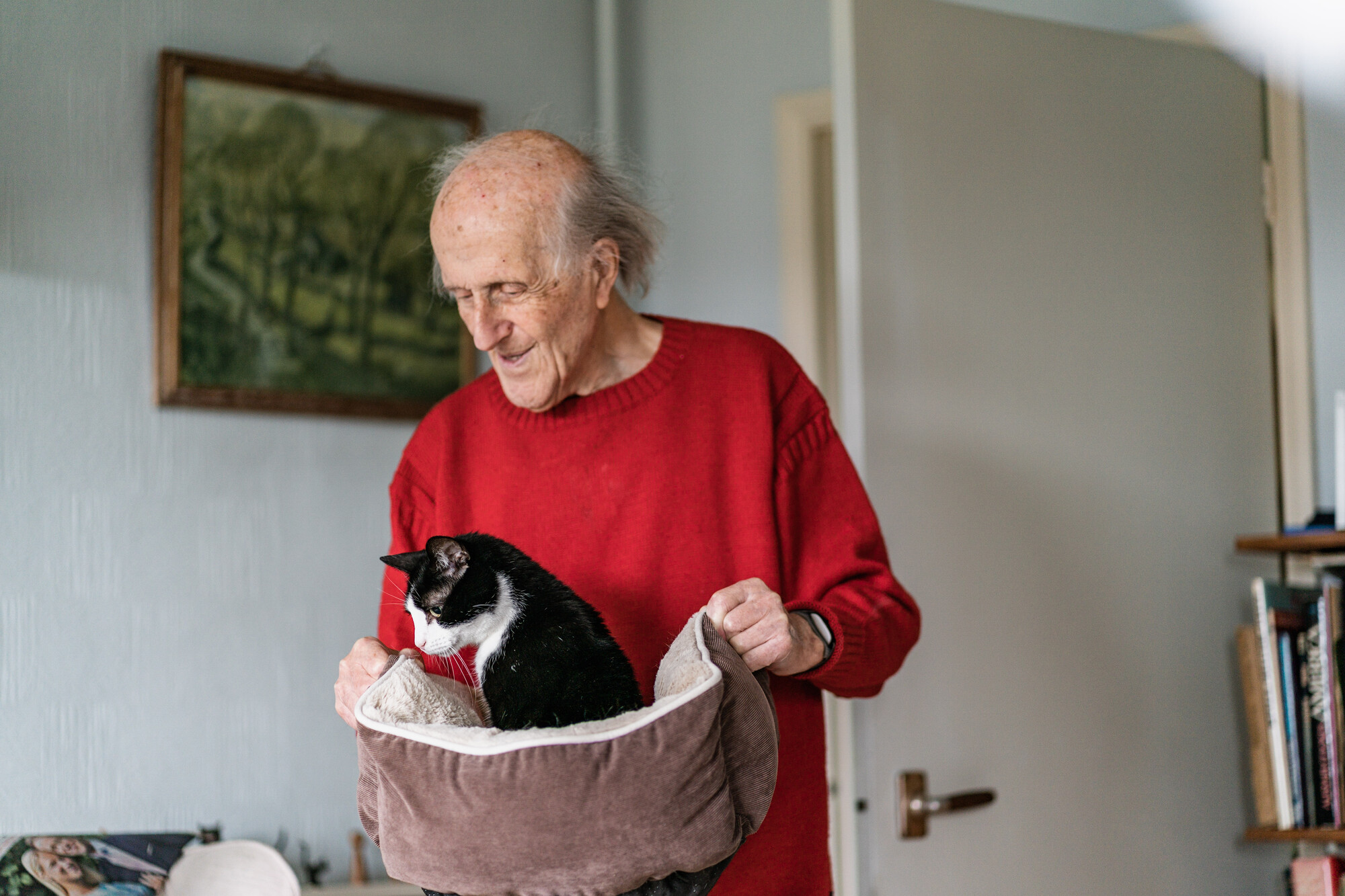 Man wearing red jumper carrying black and white in a cat bed