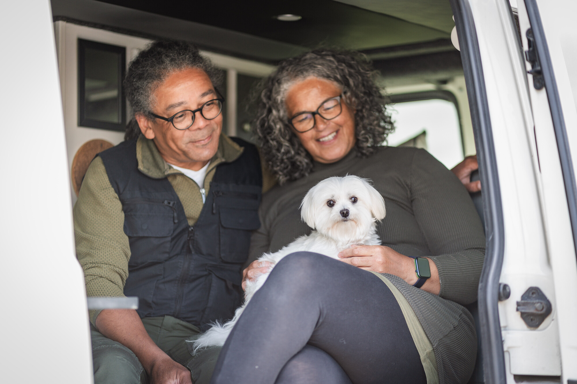A small white Maltese dog called Midge sitting in a camper van with her two owners, Eliza and Danny