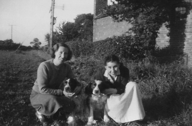 Black and white photo of Sylvia (right) and her mum (far left) with two of their dogs between them. They are in a garden with a brick building in the background.