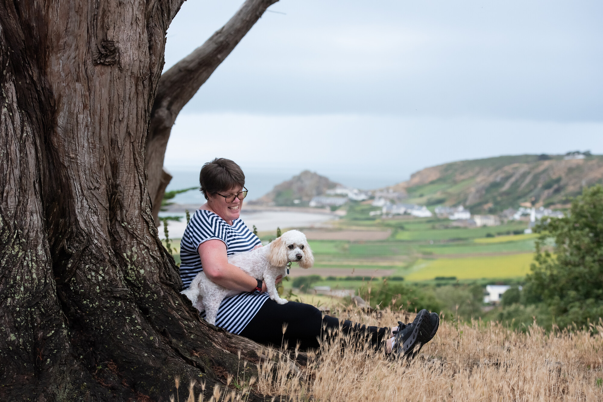 White dog Basil is perched on his owner's lap, with a beautiful green landscape in the background and the sea in the distance. His owner wears a navy and white stripey top and is leaning against a tree.