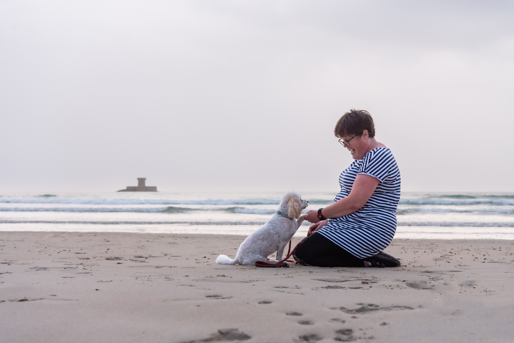 White cavapoo Basil on a beach with the sea in the background giving his owner a paw. His owner is wearing a navy and white stripey top and is looking down at him smiling.