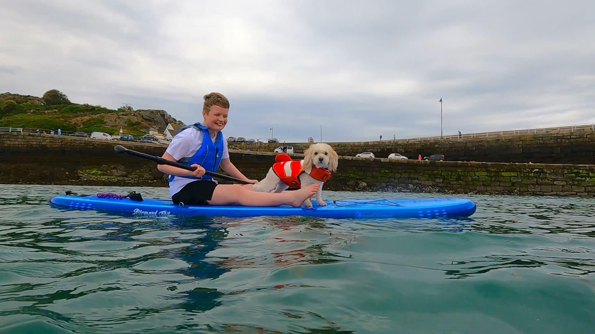 White dog Basil wearing orange life jacket on a blue paddleboard with teenager Charlotte as they paddle out to sea