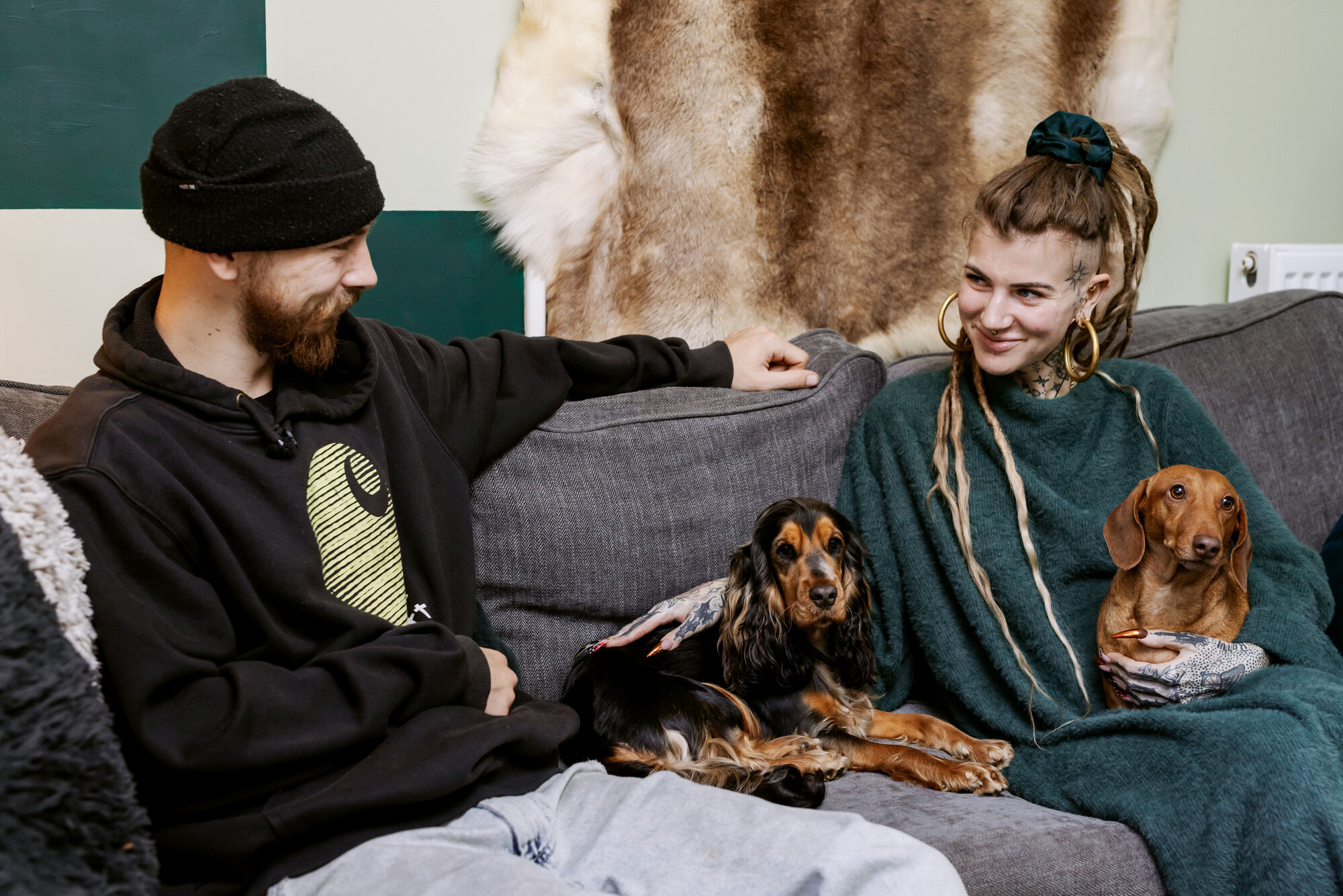 Spaniel Lilly sits on a grey sofa in between her owners and her dog best friend Barry. Her male owner is wearing a black jumper and a black hat, and her female owner is wearing a green jumper, with tan dacshund Barry on her lap