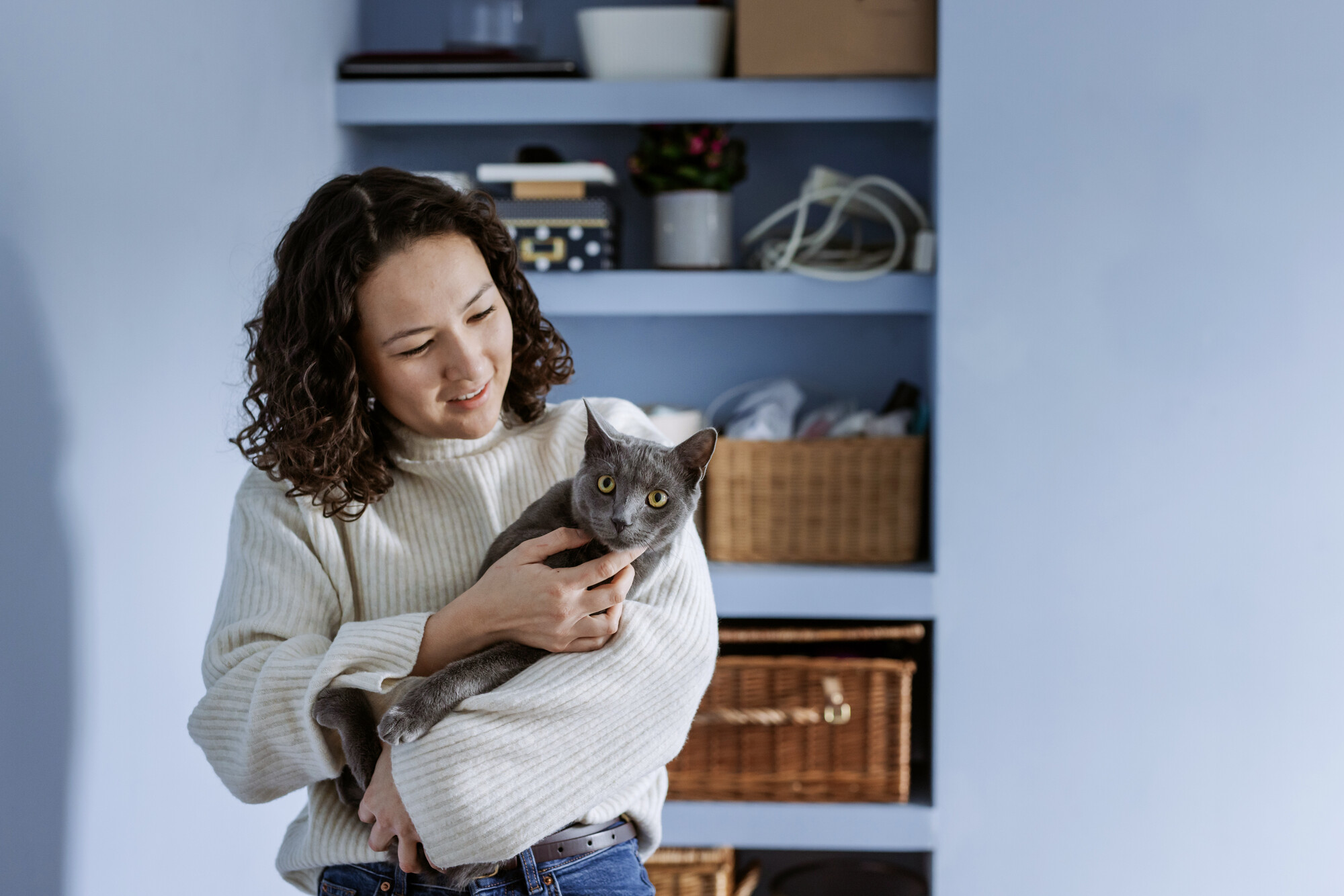 Grey cat is being held by woman in cream coloured jumper with dark curly hair. She smiles as she looks down at him. They are in front of a blue wall with shelves filled with baskets.