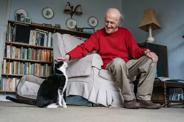 Elderly man in red jumper sits on armchair smiling and stroking black and white cat on the head
