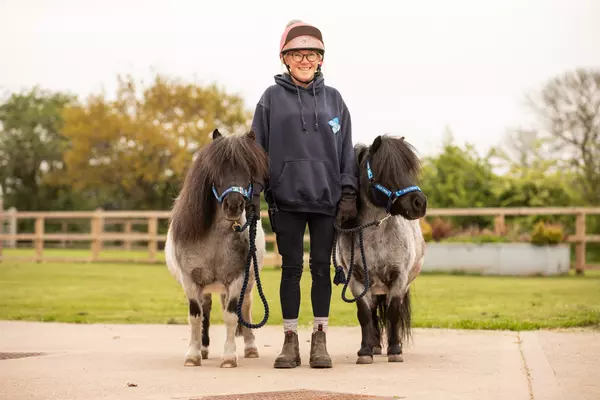 A Blue Cross groom stands between two blue roan Shetland ponies. They are outside, surrounded by greenery.