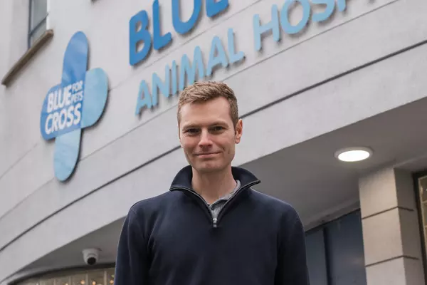 Ben the vet is a man with light skin and hair, he is smiling and wearing a blue top and is standing outside the entrance to London Victoria Hospital.