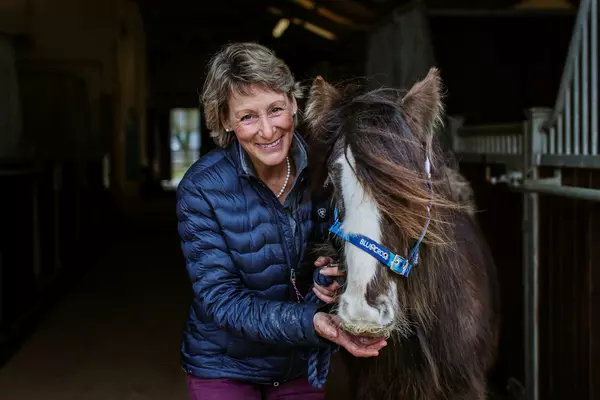 Mary King MBE DL, a lady with short , straight light hair, wearing blue and smiling, feeding a brown and white haired horse.