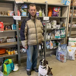 A male pet food bank owner standing in a Blue Cross pet food bank with his brown and white staffordshire bull terrier