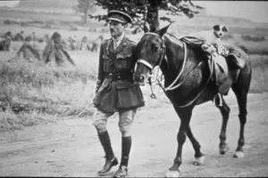 A photograph of a soldier in World War I, leading a horse tacked up. A dog is on the back of the horse.