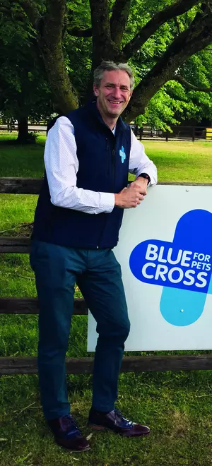 Chris Burghes leaning on a fence in front of a sign with Blue Cross's logo