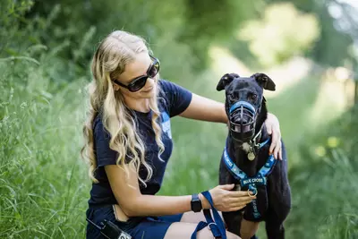 A black ex racing greyhound called Bluebell standing in a field with a blonde lady wearing sunglasses