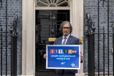 David Olusoga, a man with dark skin and grey curly hair, dressed in suit outside 10 Downing Street holding a Blue Cross end greyhound racing poster.
