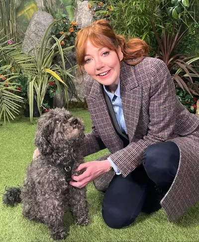 Diane Morgan, a woman with auburn hair in a ponytail, wearing a tweed coat and talking to a fluffy grey small floppy eared dog.