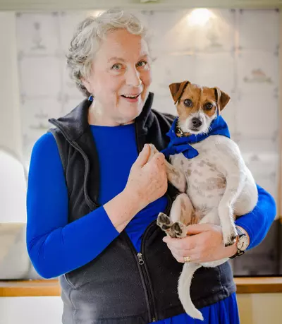 Pam Ferris is a woman with white skin and hair and is wearing a blue jumper and holding a very small dog wearing a blue scarf