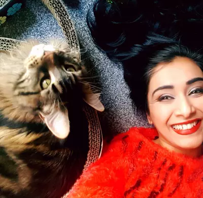 Roo Irvine, a woman with brown skin and black hair, smiling and playing with her brown and black cat.