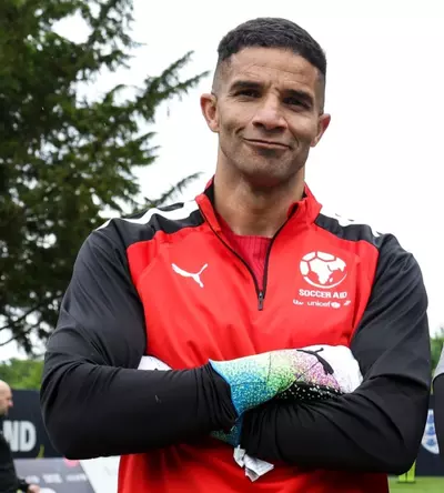 David James is a tall man with dark skin and hair and is wearing football clothing and stands with his arms crossed and is smiling.