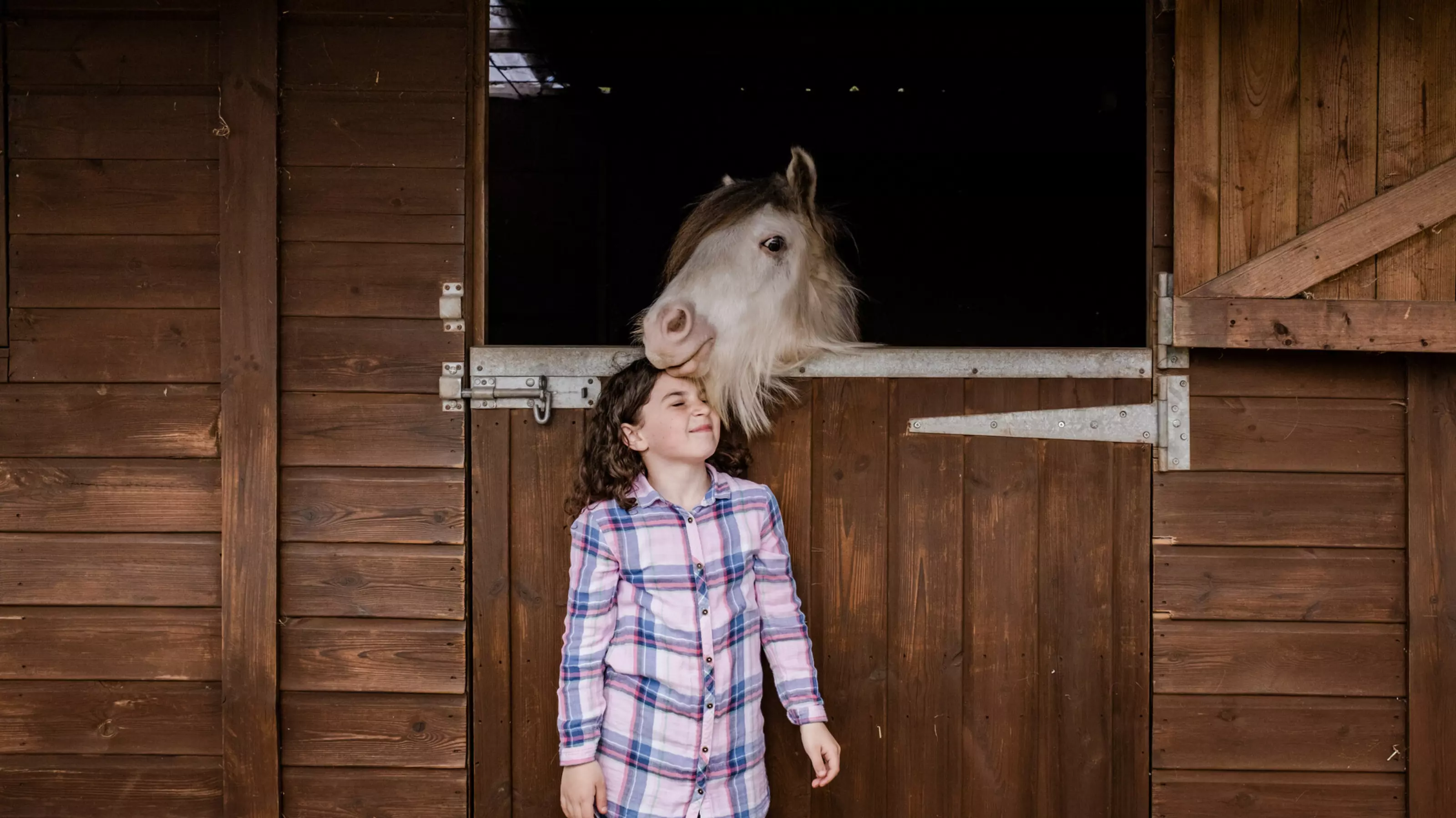 White pony Stan looking over stable door nuzzling Evie's head. Evie is wearing a pink and blue check shirt