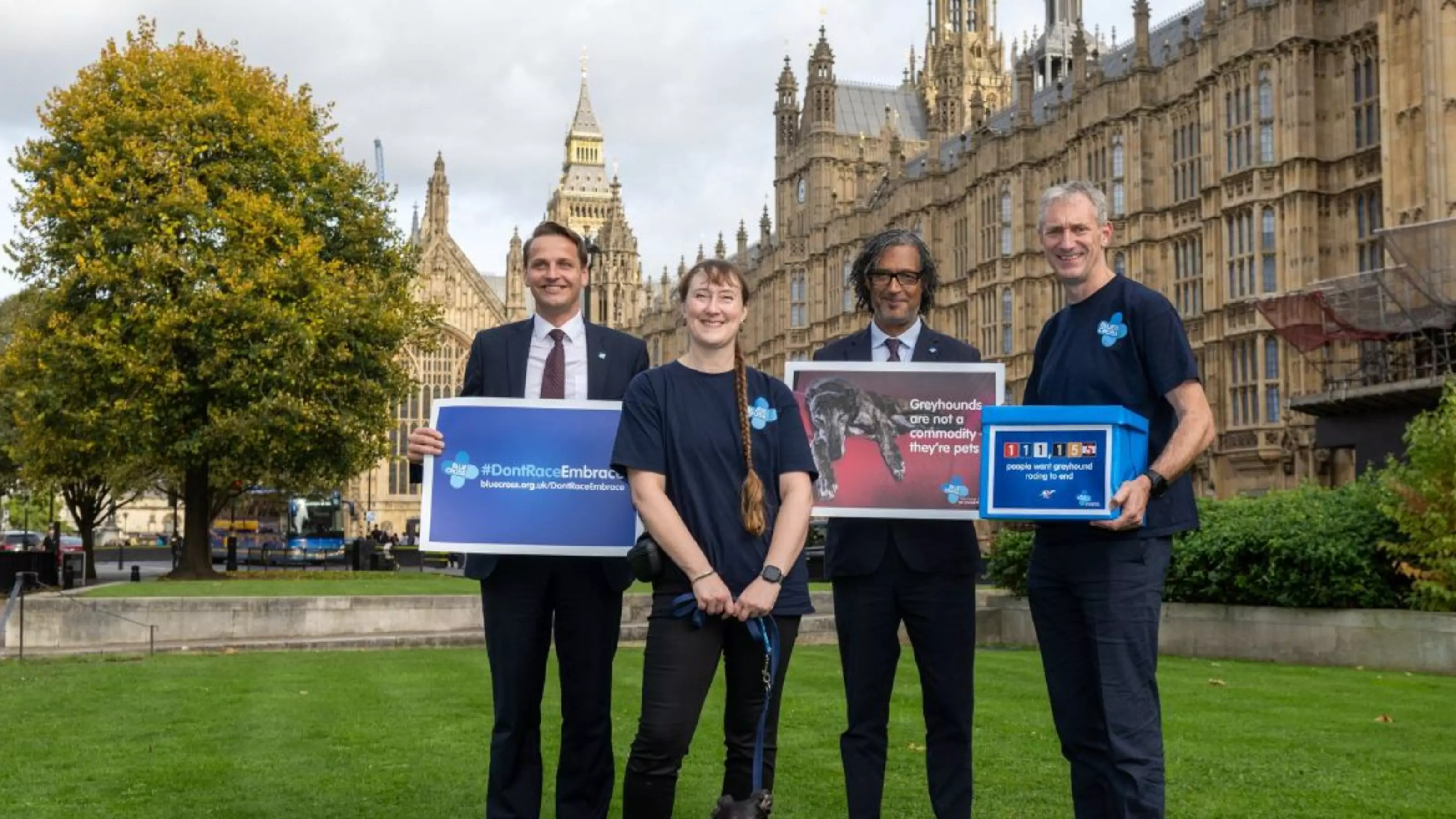 Four Blue Cross representatives outside Westminster. They're holding a box of signatures to end greyhound racing, and banners in support.