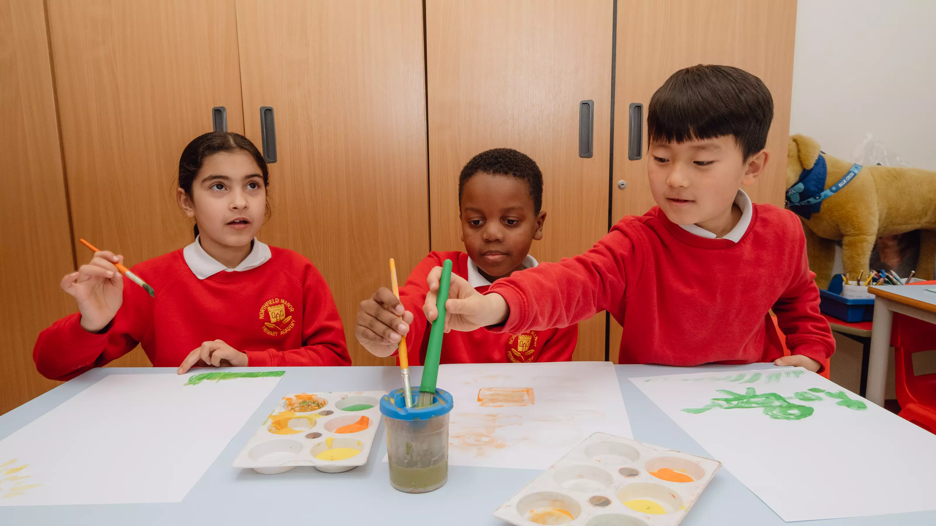 Three young primary school children painting at a desk in their classroom
