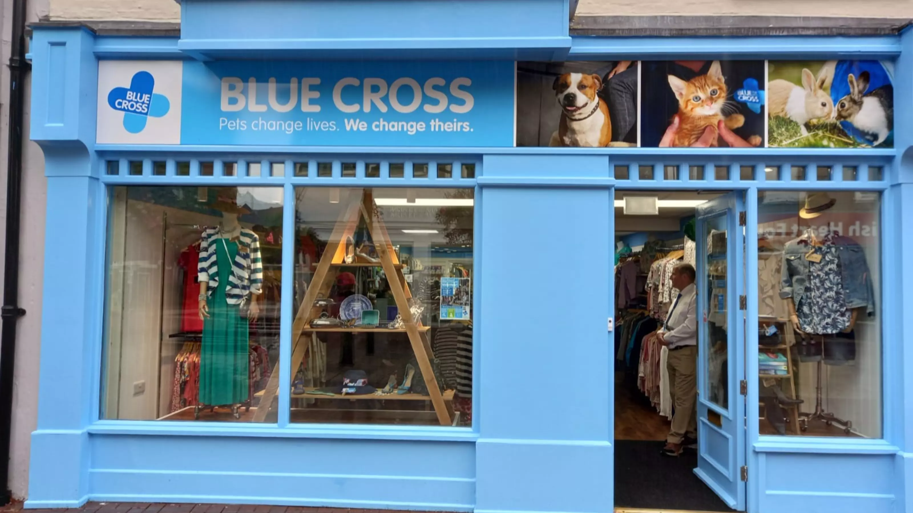 The front of a charity shop, painted blue with Blue Cross signage and neatly displayed clothing in the window.