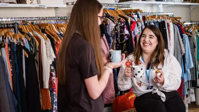 Two female volunteers in a Blue Cross charity shop having a cup of tea and smiling at each other