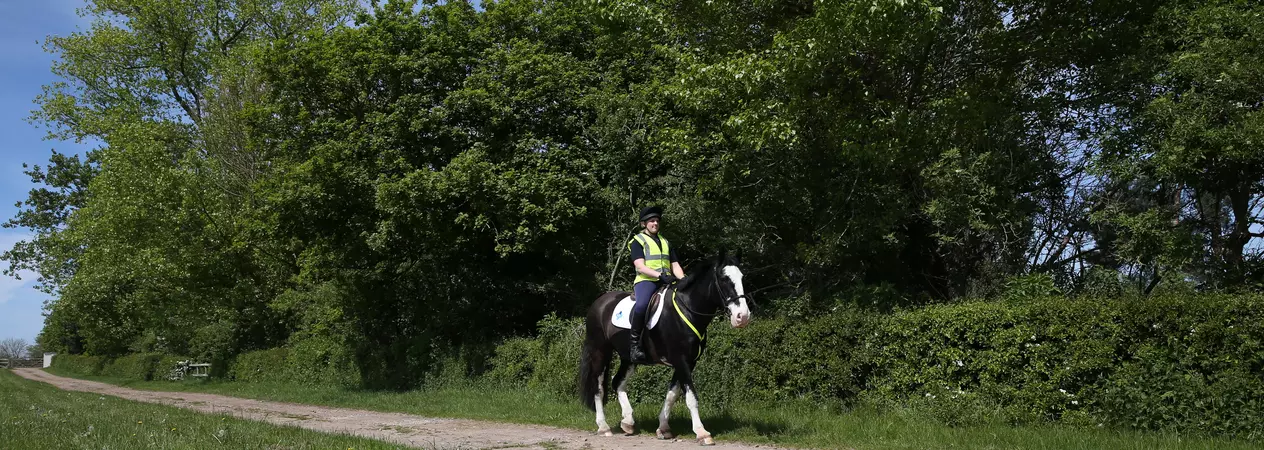 Image shows a female rider wearing a high vis jacket riding a black and white horse along a track