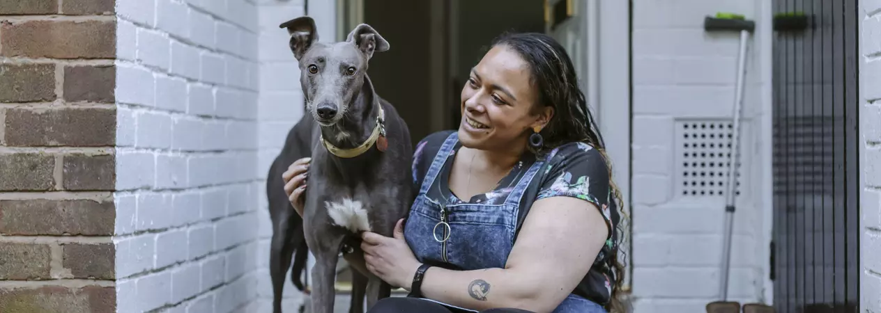 Greyhound Bleu standing on her doorstep with owner Amy sitting beside her, smiling