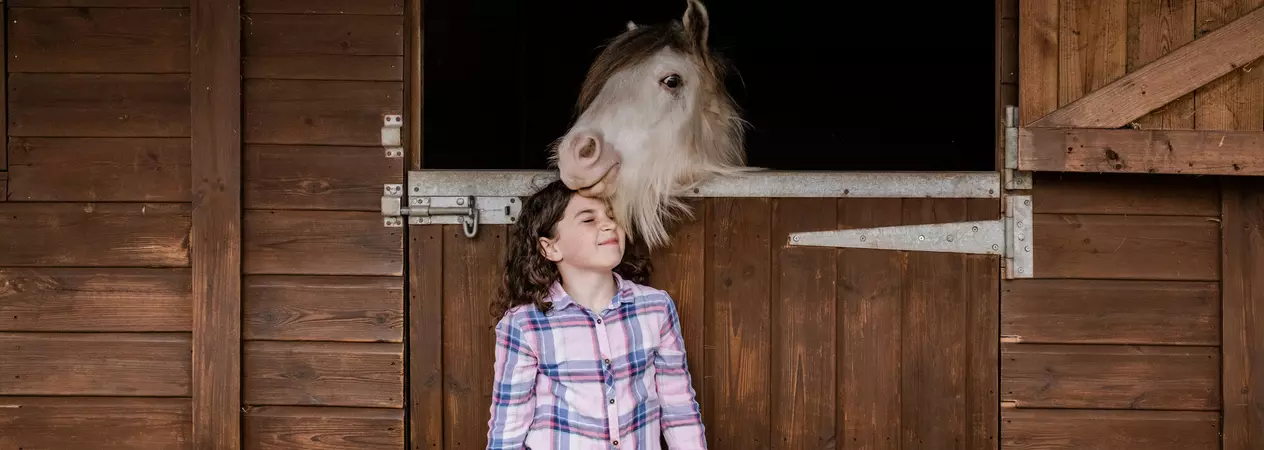 White pony Stan looking over stable door nuzzling Evie's head. Evie is wearing a pink and blue check shirt