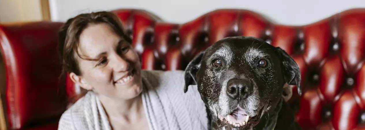 A black and white labrador cross gazes into the camera as her smiling owner wraps an arm around her
