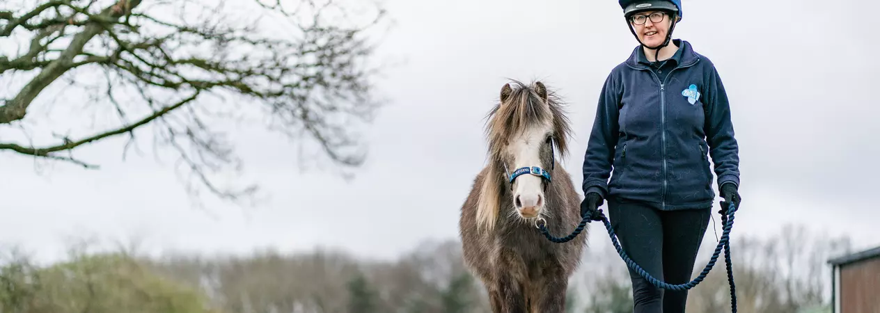 Grey pony Leo walks along a tree-lined track with his Blue Cross handler