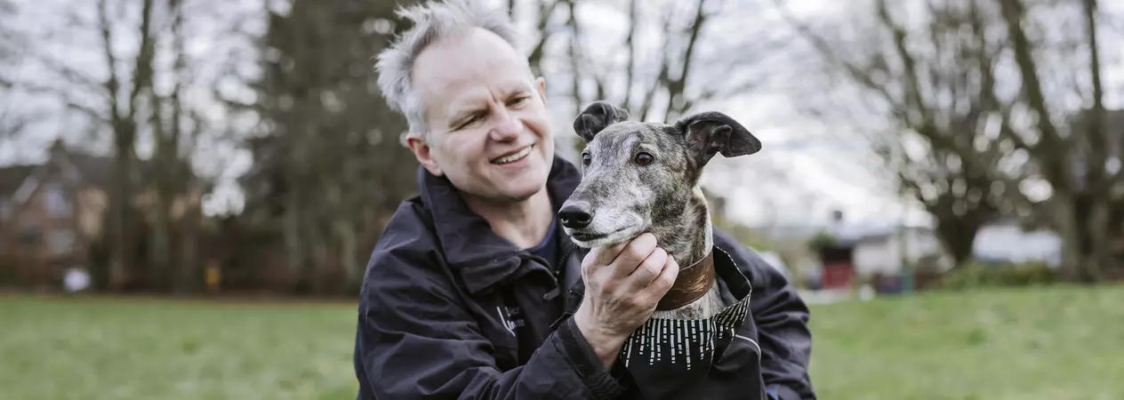 A greyhound wearing a jacket being cuddled by his owner.