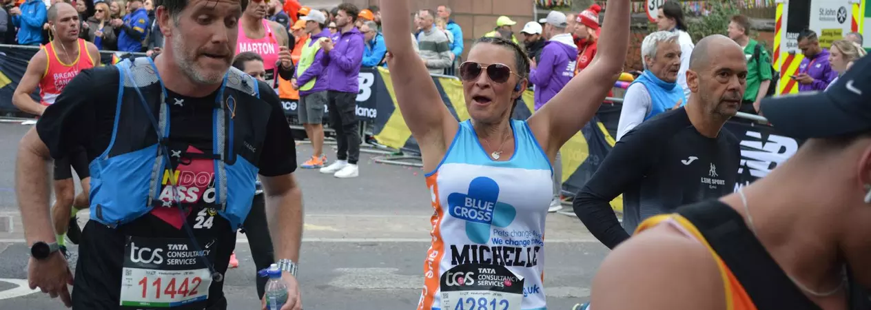 A woman participating in London Marathon, representing Blue Cross. She is holding her arms up in a cheer.