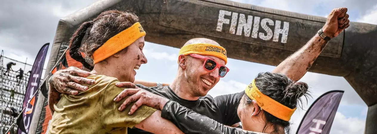 Three people celebrating at the finish line of a Tougher Mudder race. They are smiling and hugging, and covered in mud.