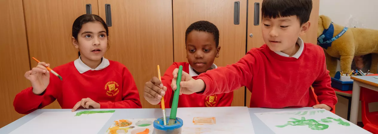 Three young primary school children painting at a desk in their classroom