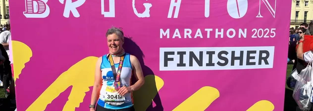 Sarah Berry, a participant of Brighton Marathon 2025, stands in front of a pink finisher sign. She wears a medal around her neck and smiles at the camera.