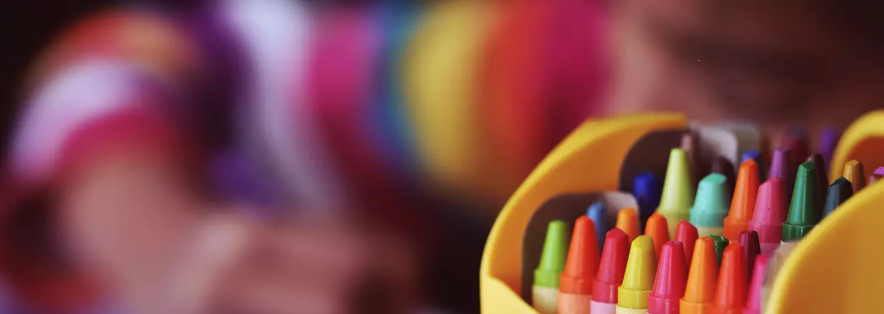 A packet of colourful crayons sits on the left of the image in focus and a blurred child can be seen behind drawing