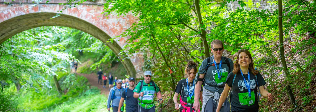 A group of walkers participate in the Windsor 50 Ultra Challenge. They are walking along a wooded path, with a bridge in the background.