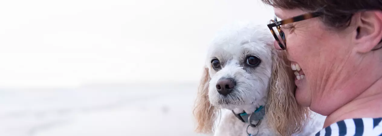 White dog Basil is held close by his owner, who smiles down at him. The beach and sea are in the background.