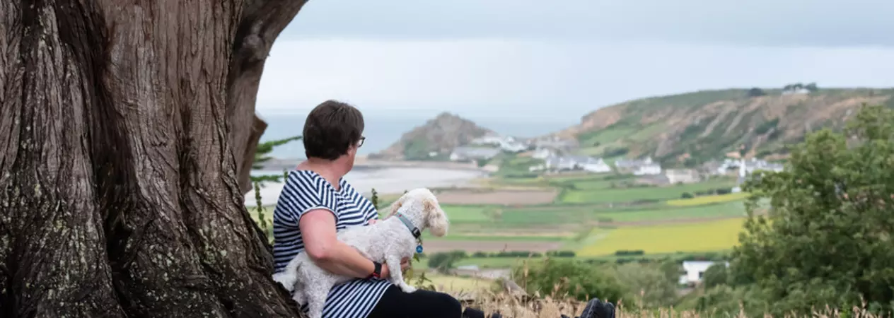 A person sitting against a large tree on a hillside, holding a small white dog while looking out over a scenic landscape of fields, hills and a distant coastline.