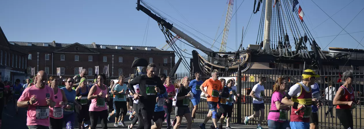 A group of runners participate in the AJ Bell Great South Run. They are running past a tall ship.
